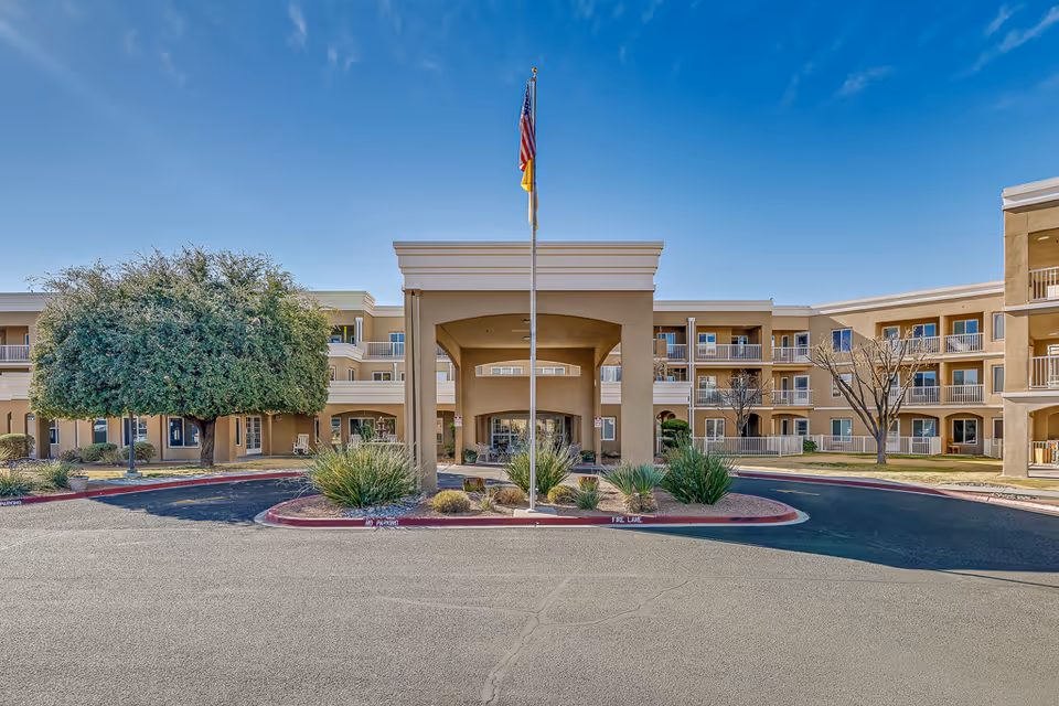 Front exterior view of Solstice Senior Living at Las Cruces building with a covered entrance, flagpole with American and New Mexico flags, landscaped plants, and a large tree on the left side under a clear blue sky.