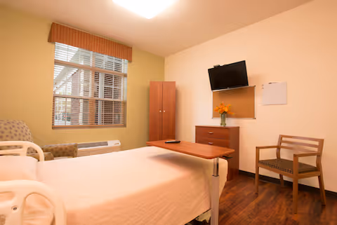 A senior living facility bedroom with a hospital-style bed covered in a light pink blanket, a wooden overbed table, a wooden chair with armrests, a wooden dresser with a vase of orange flowers, a wall-mounted flat screen TV, a corkboard, and a window with blinds and a valance.