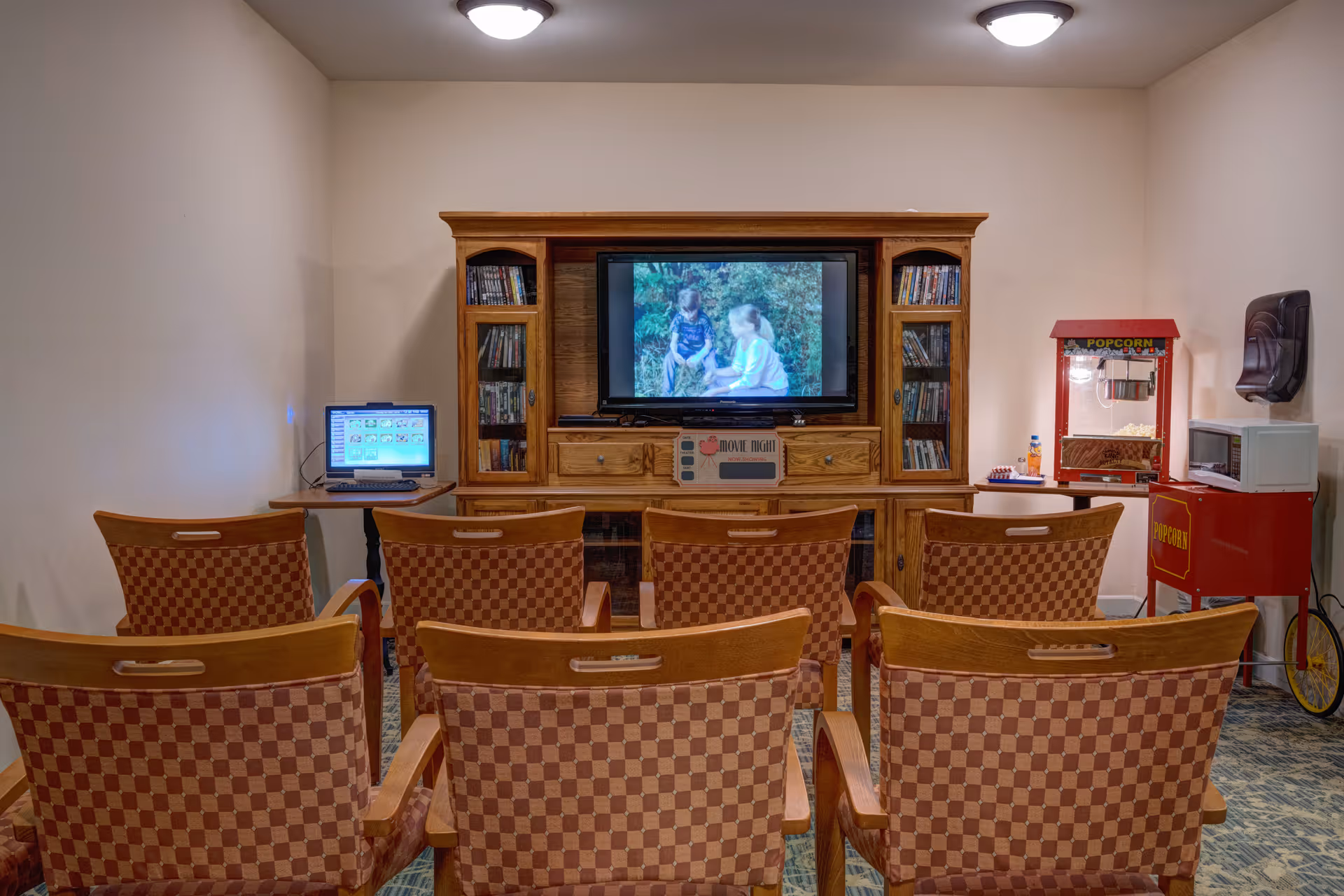 A small movie room with six checkered upholstered chairs arranged in two rows facing a wooden entertainment center with a flat-screen TV showing a video of two children. To the left of the TV is a small table with a laptop, and to the right is a red popcorn machine and a microwave on a small table. The room has beige walls and ceiling lights.