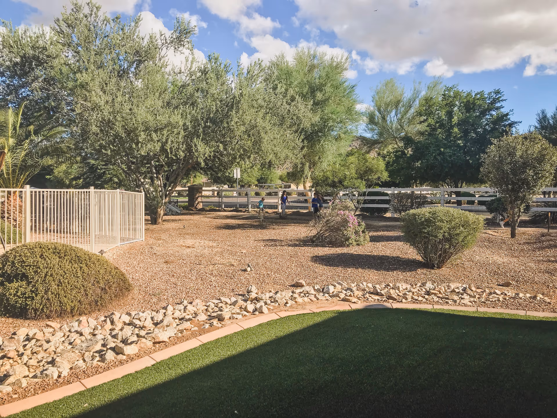 Landscaped outdoor courtyard with gravel beds, scattered shrubs and trees, a white fence, and a small patch of green lawn under a partly cloudy sky.