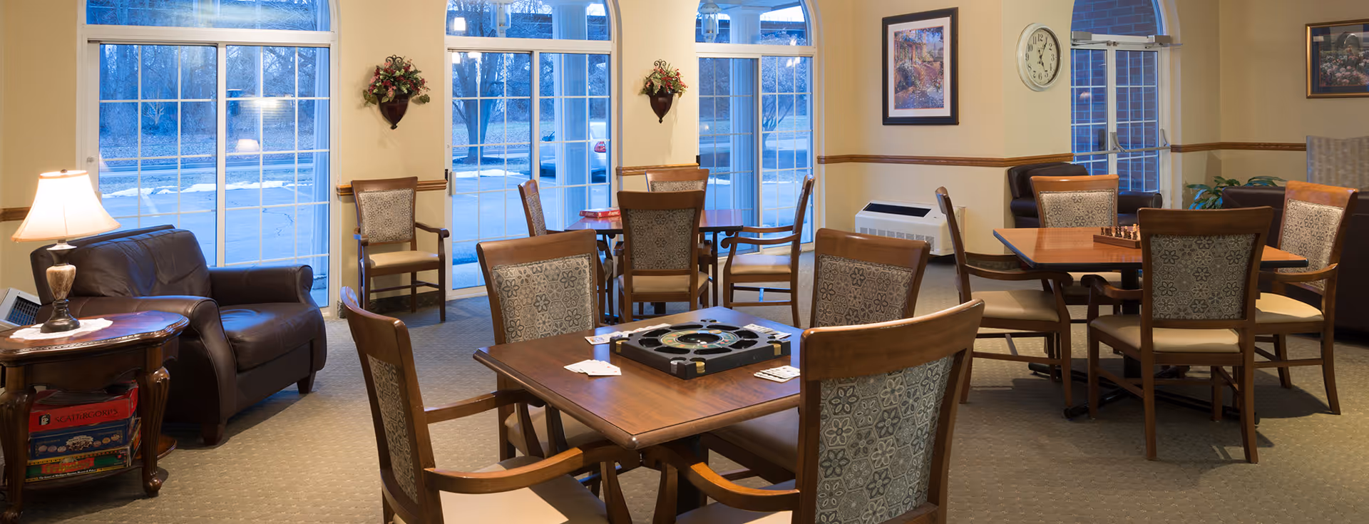 A well-lit common area with multiple wooden tables and chairs arranged for games and social activities. The room features large windows with a view of the outside, a leather armchair next to a small table with a lamp, and framed artwork on the walls. Board games and playing cards are visible on the tables.