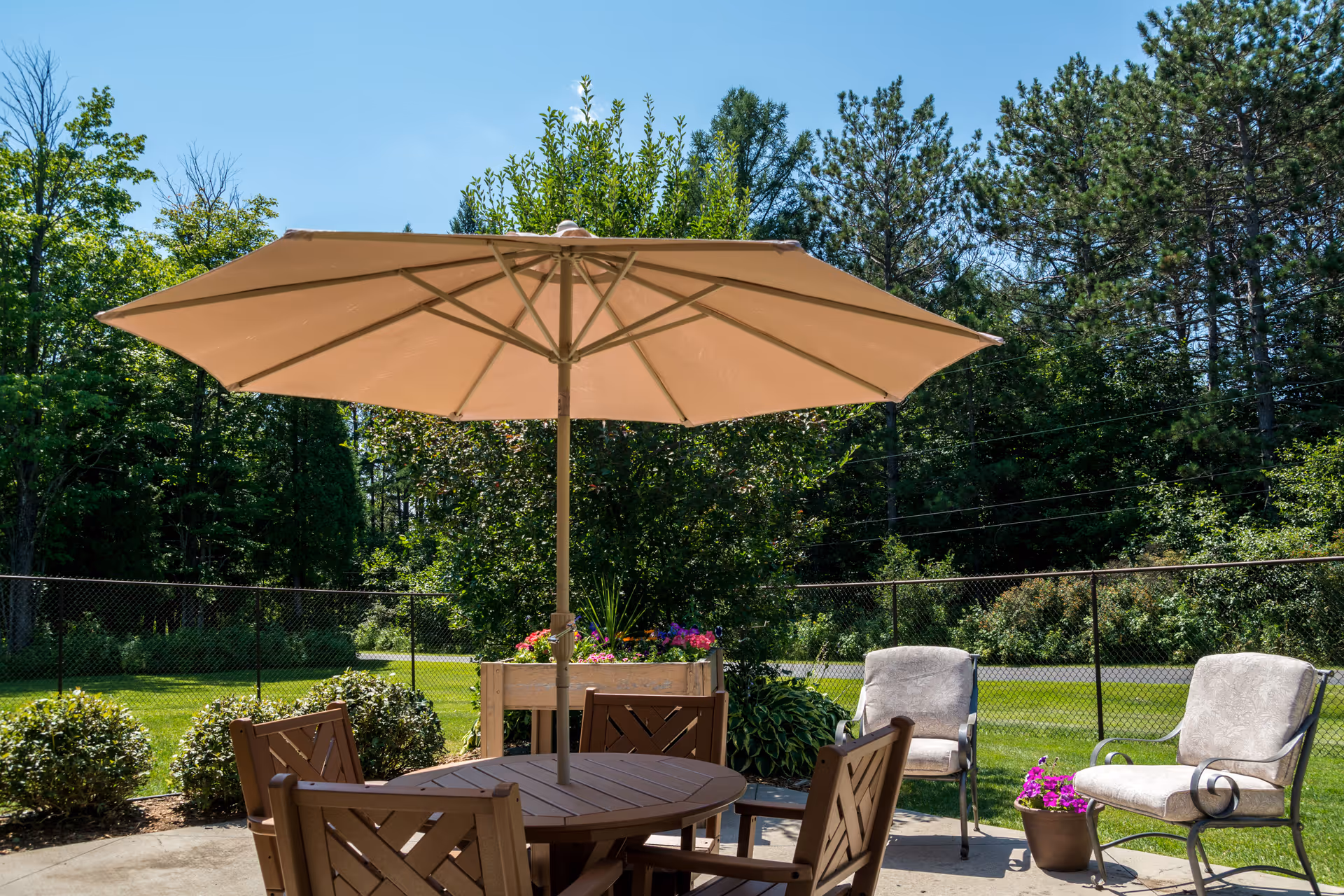 Patio seating area with a round table, large umbrella, chairs, and potted plants beside a fenced lawn and trees.