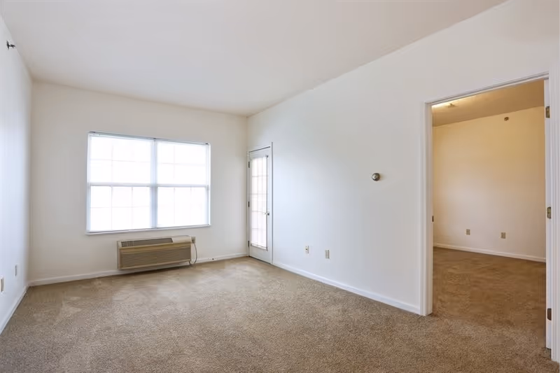 Empty living room with a large window, wall air-conditioning unit, beige carpet, and a doorway to an adjacent room.