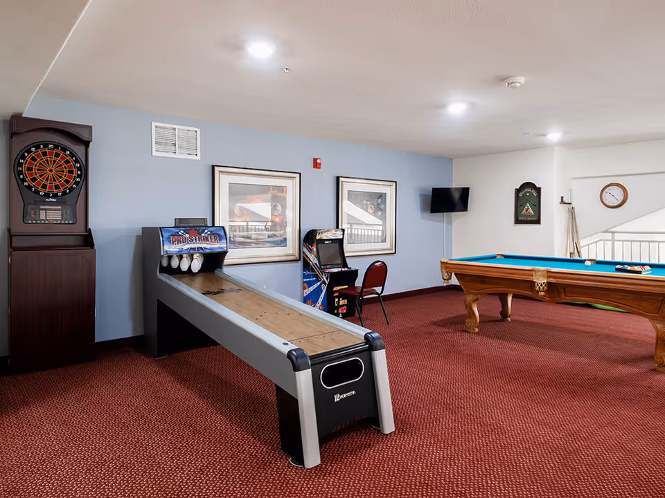 A game room with a dartboard, a Pro Striker bowling game, an arcade machine, and a pool table with balls set up. The room has red carpet, light blue and white walls, framed pictures, a wall-mounted TV, and a clock on the wall.