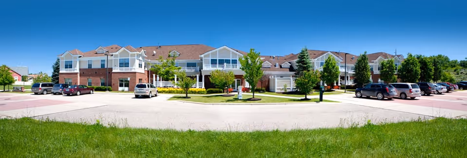 Wide panoramic view of The Cortland Howell senior living facility showing a large two-story building with a brick and white exterior, multiple windows, and a central entrance. The building is surrounded by a parking lot with several parked cars and landscaped greenery including trees and grass under a clear blue sky.