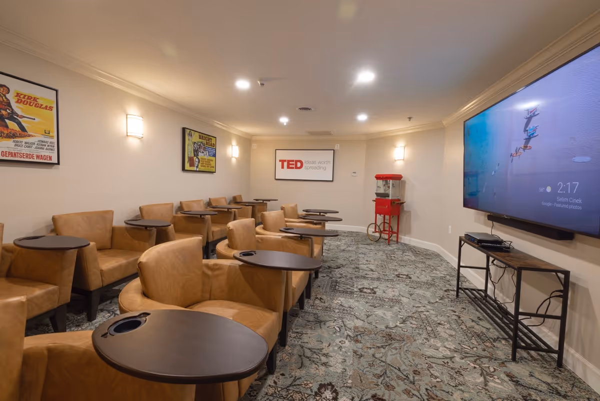 A cozy media room with multiple tan leather armchairs, each equipped with a small attached dark wooden table. The room has a patterned carpet and beige walls adorned with framed posters and a TED Ideas Worth Spreading sign. A large flat-screen TV is mounted on the right wall, and a vintage-style red popcorn machine stands in the corner.