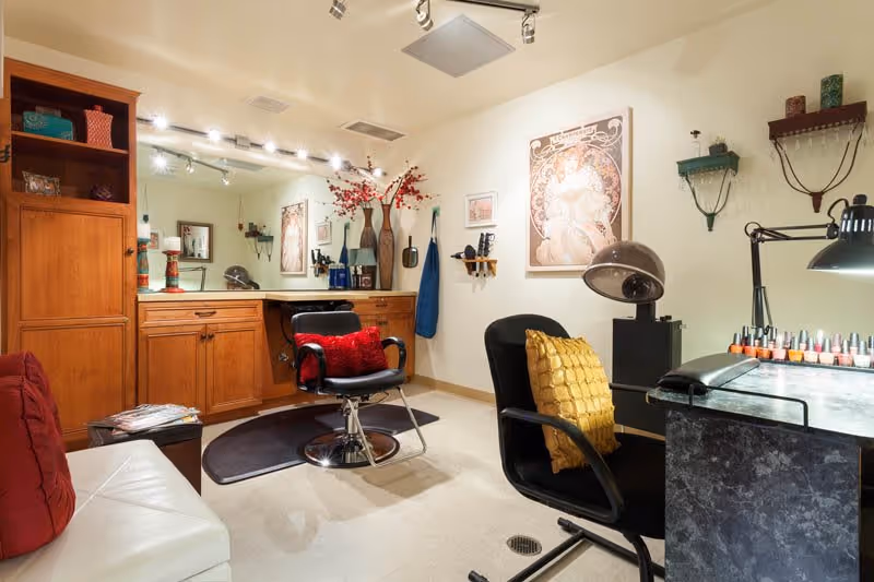 Interior view of a salon area with a styling chair in front of a large mirror and wooden cabinets. There is a white couch with a red pillow, a black chair with a yellow pillow, a nail polish station with various nail polish bottles, and a vintage hair dryer. The walls are decorated with framed artwork and shelves holding decorative items.