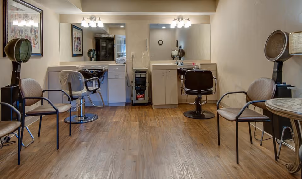 Interior view of a hair salon area in a senior living facility with two salon chairs in front of mirrors and sinks, surrounded by waiting chairs and hair drying stations. The room has wooden flooring and neutral-colored walls with framed artwork.
