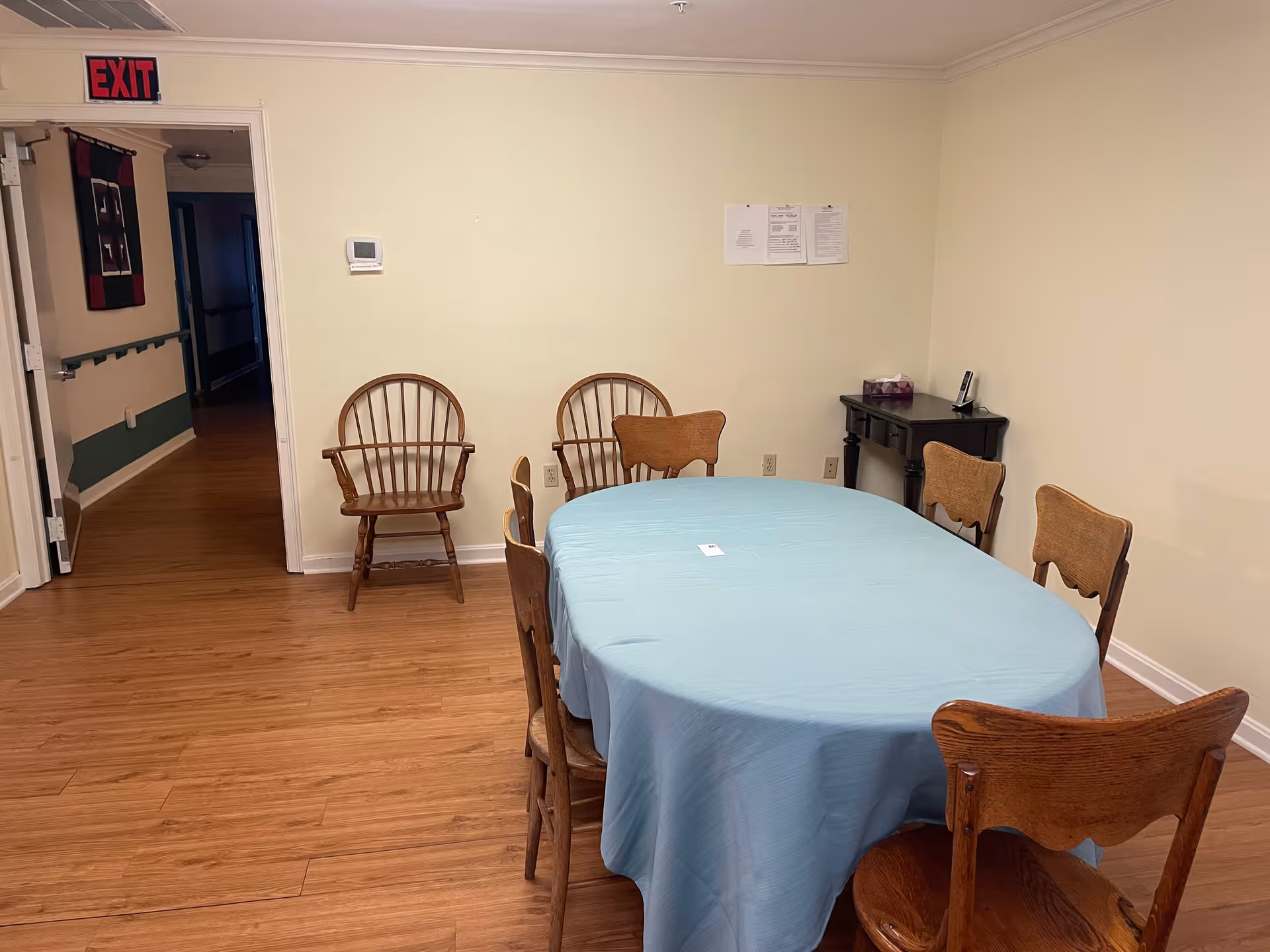 Small dining room with an oval table covered by a light blue tablecloth surrounded by wooden chairs, hardwood floor and an exit sign visible in the hallway.