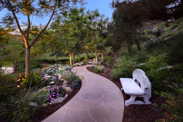 Curving stone walkway through a landscaped garden with trees, flowers, and a white stone bench.