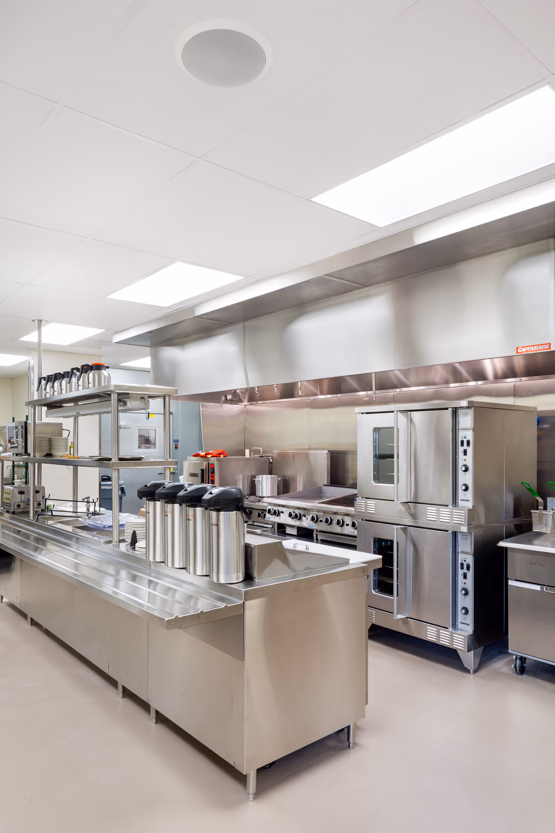 A clean, modern commercial kitchen with stainless steel appliances and countertops. The kitchen features multiple ovens, a large stove with multiple burners, a stainless steel ventilation hood, and several coffee dispensers on the counter. The space is well-lit with bright overhead lights and has a polished floor.
