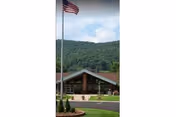 Exterior view of a single-story building with a peaked roof, large windows, and a flagpole with an American flag in front. The building is surrounded by a well-maintained lawn and landscaping, with green hills in the background under a partly cloudy sky.