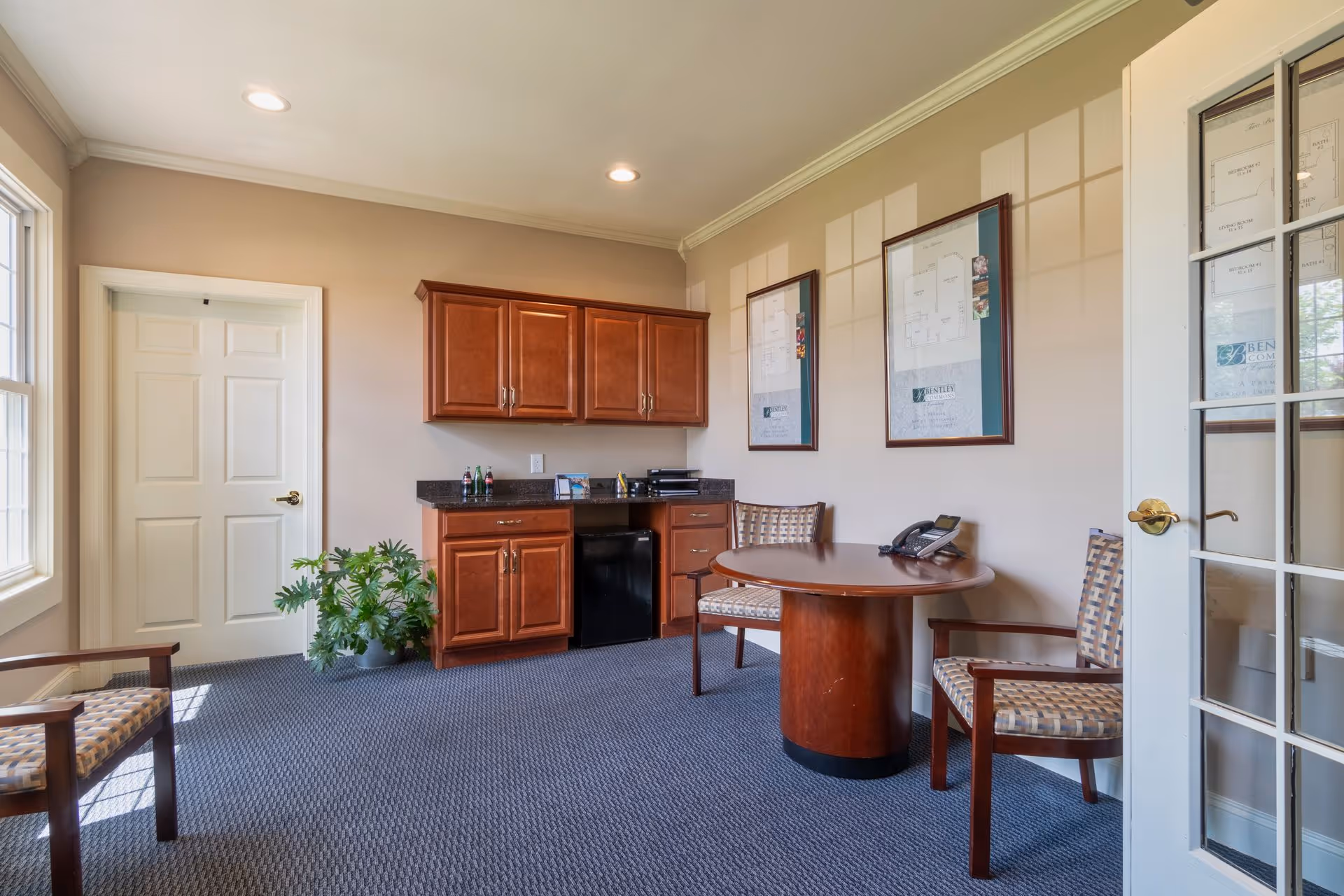 A small office or meeting room with a round wooden table and three patterned chairs. There is a wooden cabinet with a black countertop and a small black refrigerator underneath. Two framed floor plans are hung on the beige wall. A potted plant is placed near a white door, and a glass-paneled door is partially open on the right side.