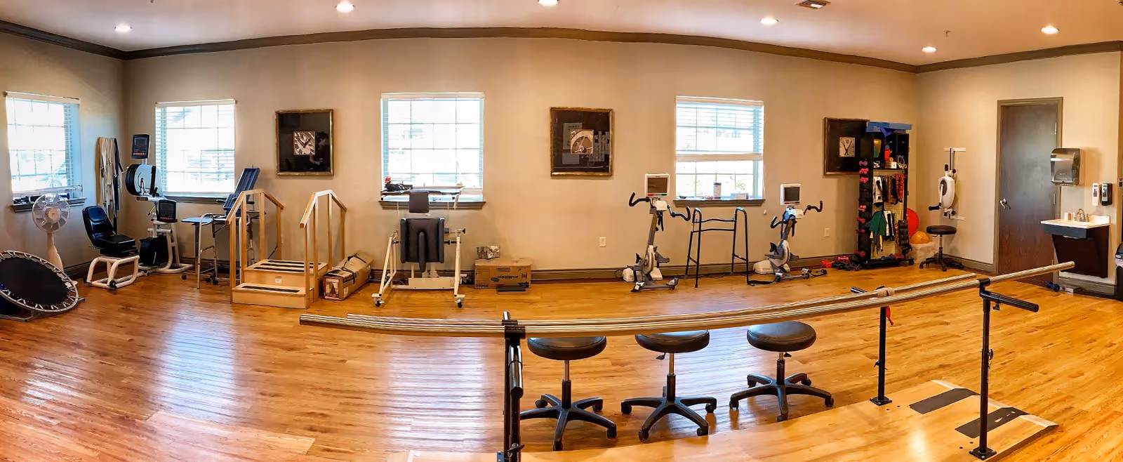 A spacious rehabilitation or physical therapy room with wooden floors and beige walls. The room contains various exercise and therapy equipment including stationary bikes, parallel bars, a small trampoline, a walker, and therapy stairs. There are three stools in front of the parallel bars, windows letting in natural light, framed pictures on the walls, and a sink with a paper towel dispenser near a closed door.