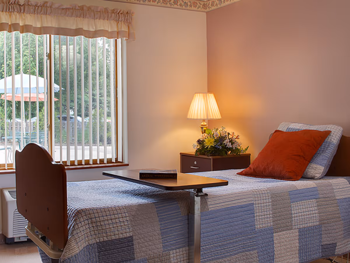 A single bed with a patchwork quilt and a red pillow in a softly lit room. Next to the bed is a small nightstand with a lamp and a floral arrangement. A window with vertical blinds shows an outdoor patio area with umbrellas and greenery.