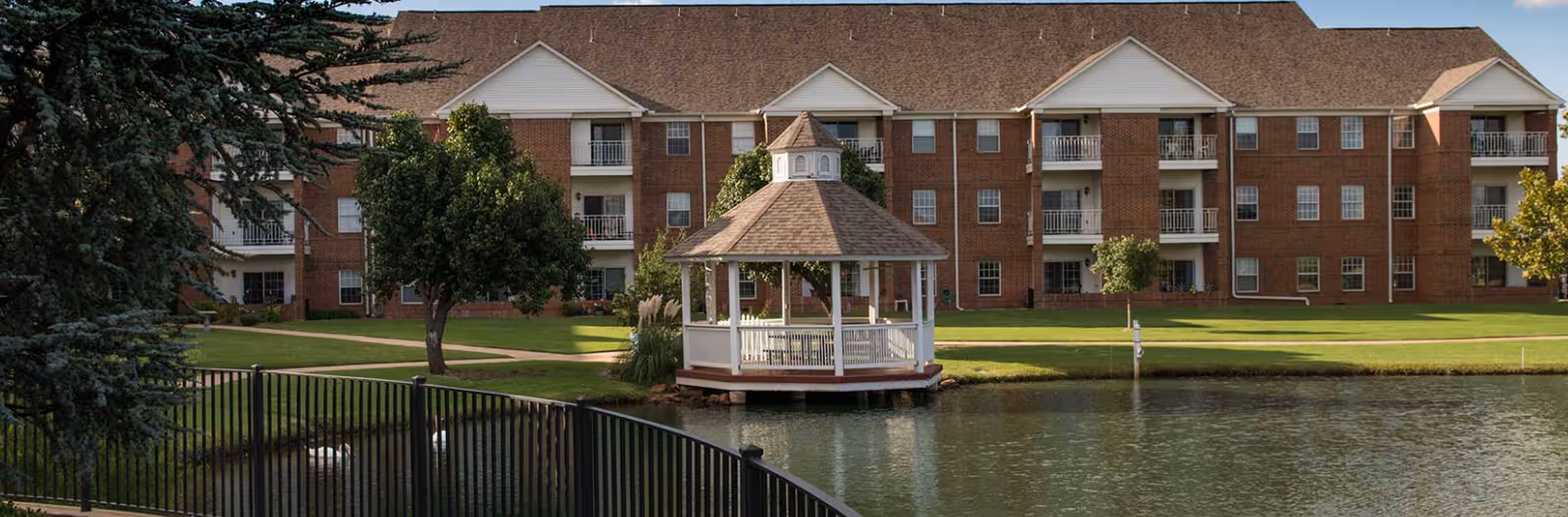 Brick multi-story senior living building with balconies overlooking a pond and a white gazebo, with a black fence and trees in the foreground.