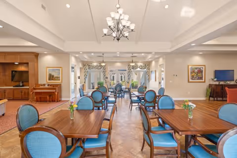 A spacious and well-lit dining room with multiple wooden tables and blue cushioned chairs. Each table has a small vase with flowers. The room features a chandelier, recessed lighting, framed artwork on the walls, and large windows with curtains allowing natural light to enter. There is a piano and a TV on wooden cabinets along the walls.