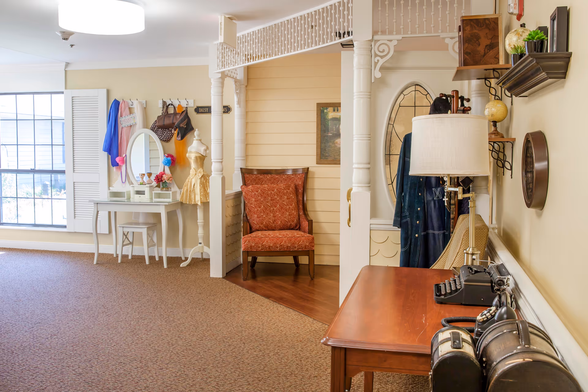 A cozy interior space featuring a vintage-style wooden desk with an old-fashioned typewriter and rotary phone, a lamp, and decorative items on shelves. To the left, there is a white vanity table with a round mirror, a stool, and various accessories hanging on hooks above it. A red upholstered chair is placed in a small alcove with wooden paneling and decorative white columns. A large window with white shutters allows natural light into the room.