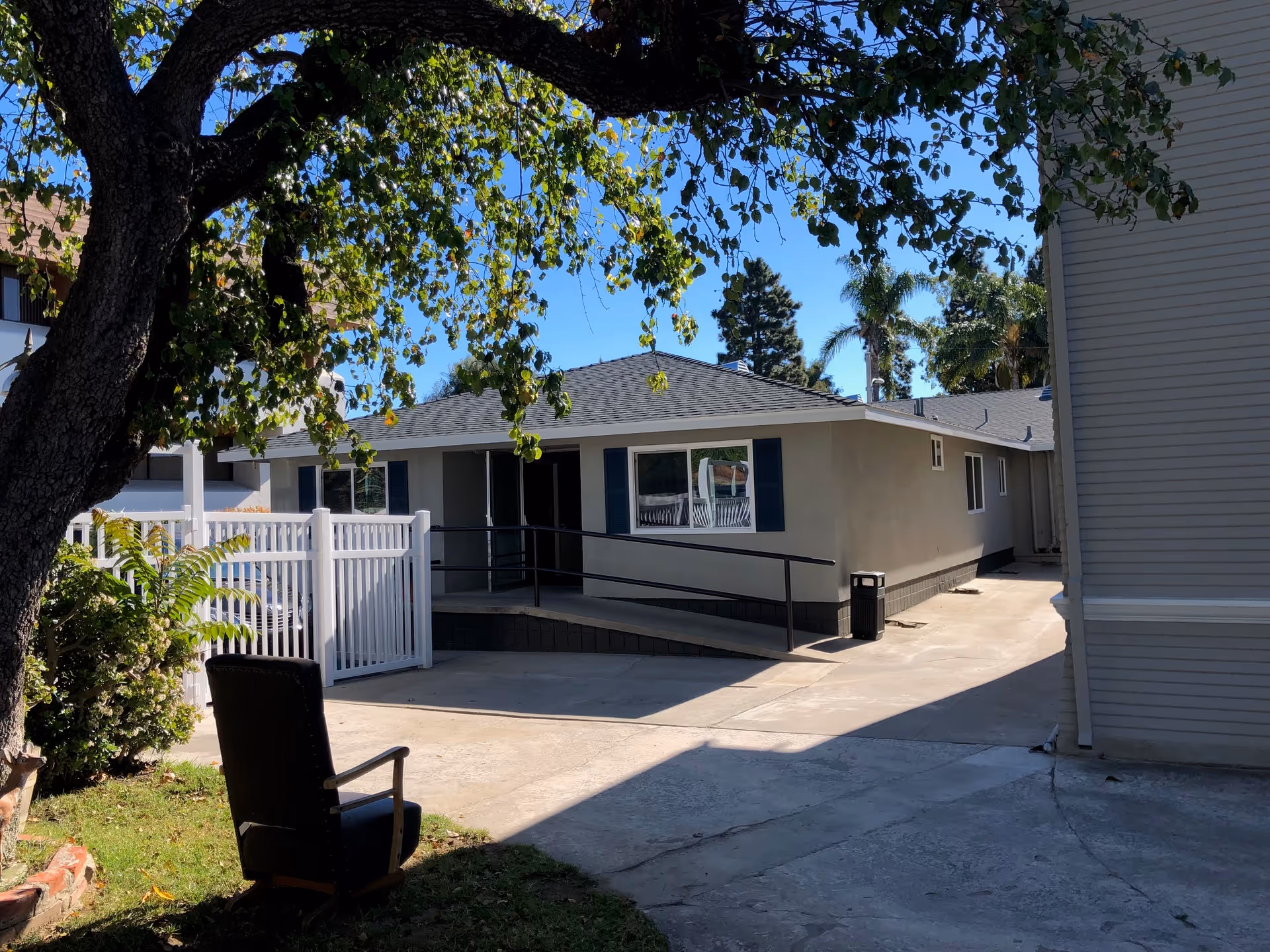 Outdoor view of a single-story building with a wheelchair ramp and a white fence. There is a tree with green leaves in the foreground and a black chair on the grass near the tree. The sky is clear and blue.