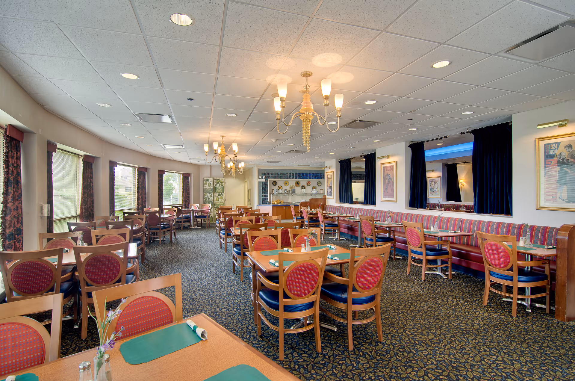 A spacious dining room with multiple wooden tables and chairs arranged neatly. The chairs have red cushioned backs and blue seats. The room features large windows with floral curtains on one side, allowing natural light to enter. There are chandeliers hanging from the ceiling, and the walls are decorated with framed pictures and mirrors with dark blue curtains. The carpet has a patterned design, and the overall ambiance is warm and inviting.