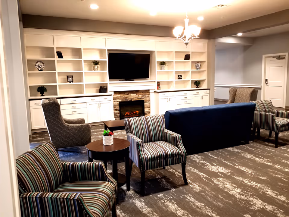 A cozy living room area in Corn Heritage Village featuring a blue sofa, two striped armchairs, and two gray armchairs arranged around a small round wooden table with a small plant. The room has a built-in white shelving unit with cabinets, a flat-screen TV mounted above a stone fireplace, and soft overhead lighting from a chandelier.