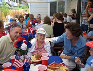 A group of elderly people and caregivers gathered around tables outdoors, enjoying a meal with red, white, and blue decorations including flowers and small American flags.