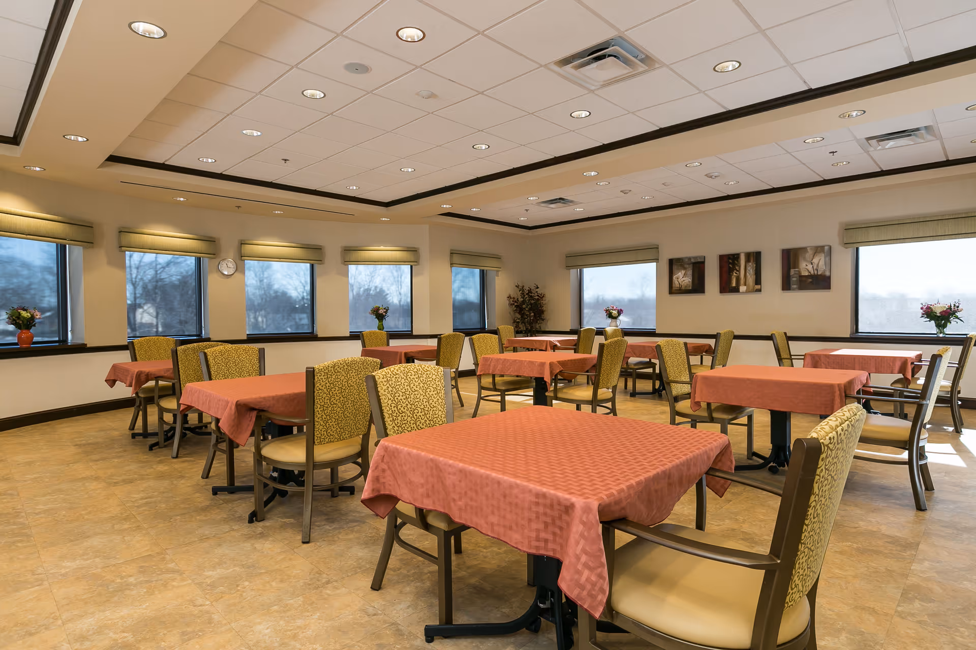 A bright dining room with multiple tables covered in red tablecloths and surrounded by cushioned chairs. The room has large windows with green valances, allowing natural light to fill the space. There are flower arrangements on the windowsills and framed artwork on the walls.