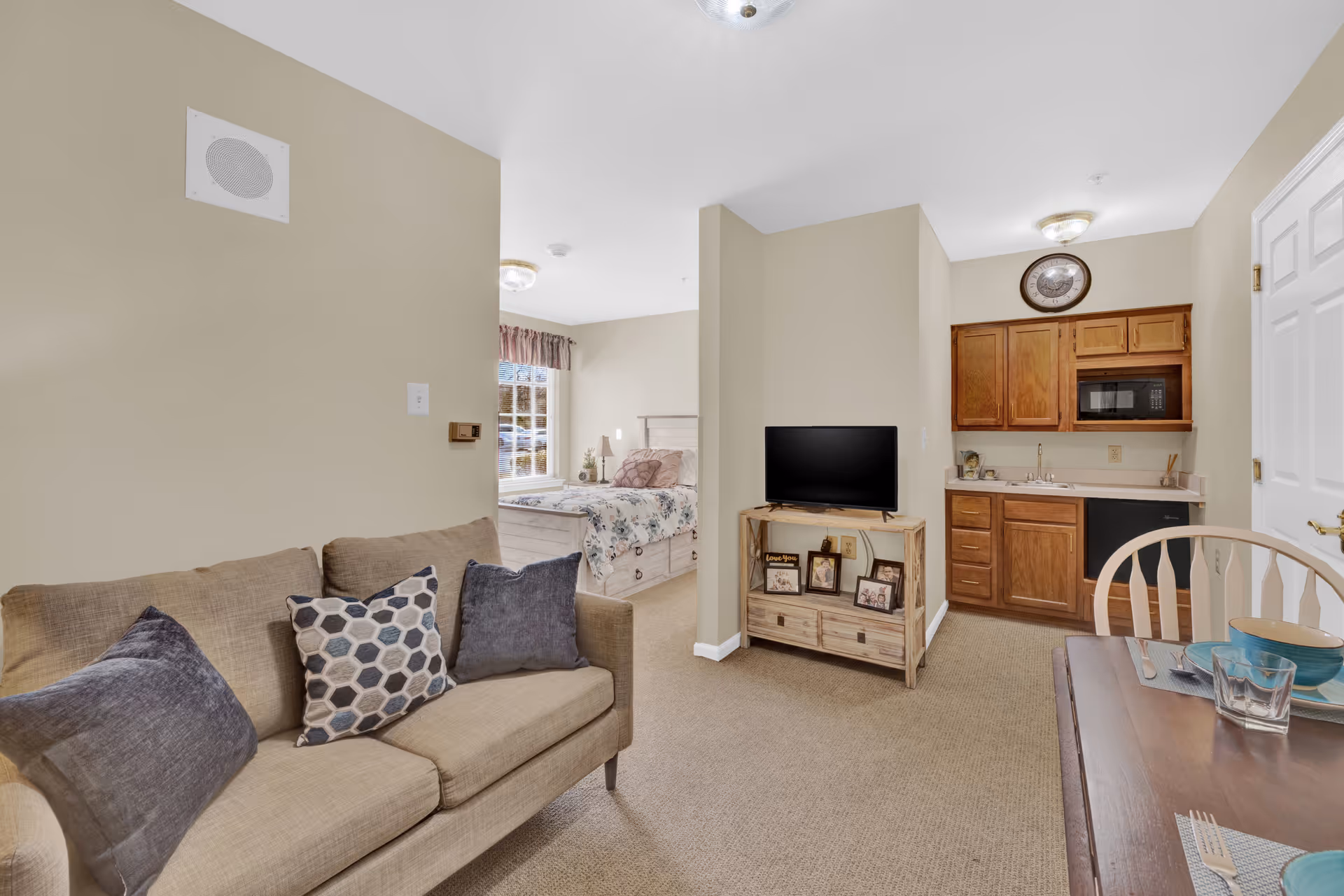 Interior view of a senior living facility apartment at Juniper Village at Williamstown featuring a beige sofa with three pillows, a small wooden TV stand with a flat-screen TV and framed photos, a kitchenette with wooden cabinets, a microwave, and a sink, a dining table set with blue bowls and glasses, and a bedroom area with a bed covered in floral bedding visible through an open doorway.
