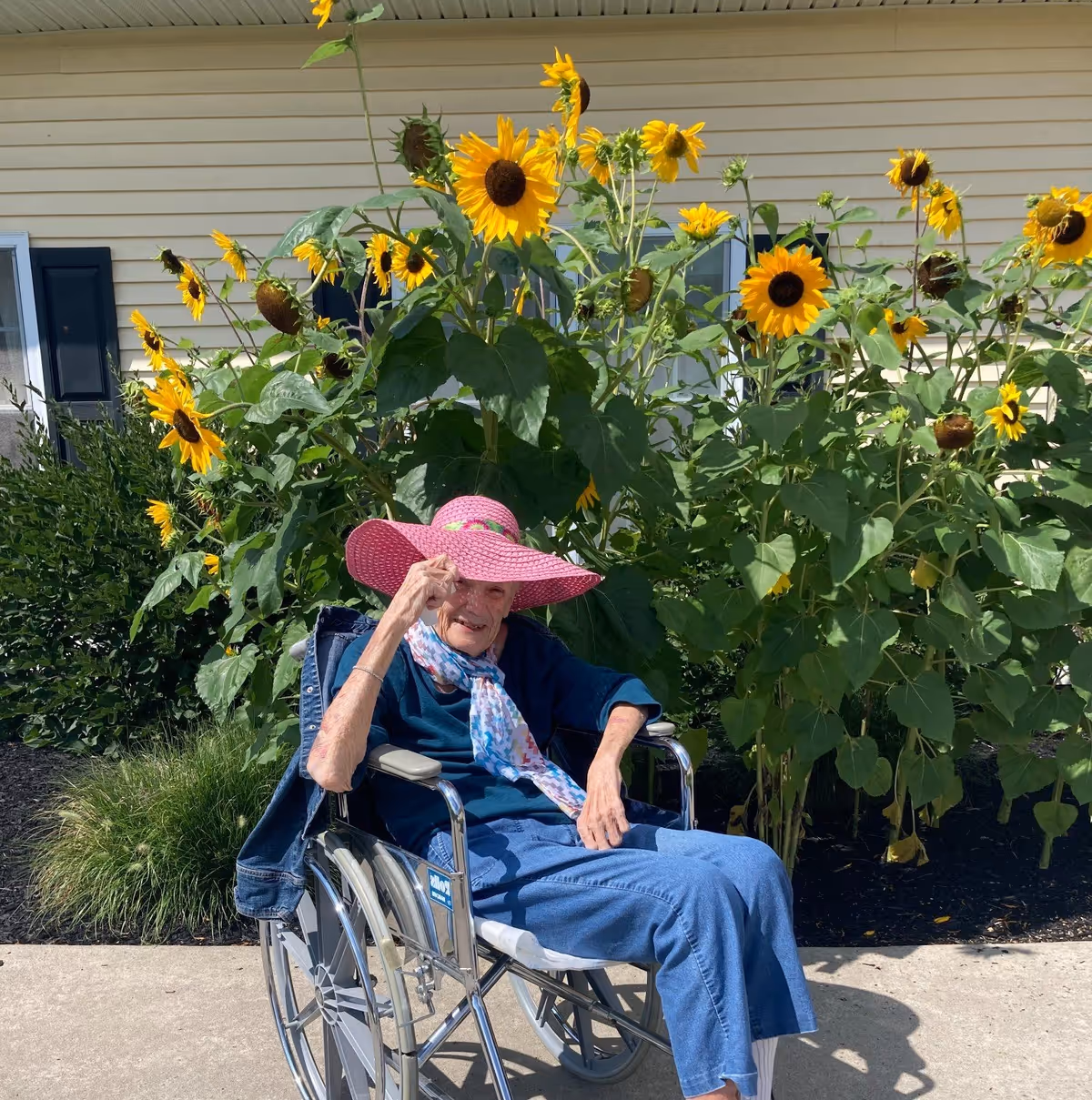 An elderly woman sitting in a wheelchair outdoors in front of tall sunflowers. She is wearing a large pink sunhat, a blue long-sleeve shirt, blue pants, white socks, and a colorful scarf. A denim jacket is draped over the back of the wheelchair. The background shows a beige building with black shutters.