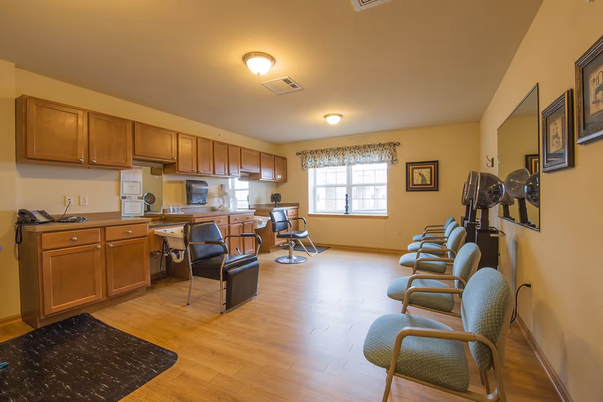 Interior view of a salon area in a senior living facility with wooden cabinets along one wall, two black salon chairs, a row of six light blue cushioned chairs with armrests along the opposite wall, and two hair dryers mounted on stands. The room has wood flooring, beige walls, a large window with a floral valance, and framed artwork on the walls.