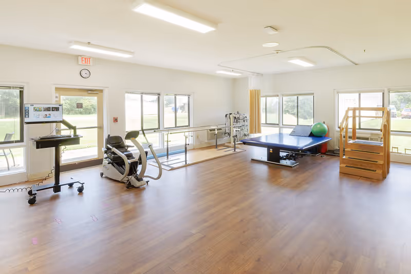A spacious physical therapy room with wooden flooring, large windows letting in natural light, a stationary exercise bike, parallel bars for walking exercises, a padded therapy table with an exercise ball, and wooden stairs for rehabilitation exercises.