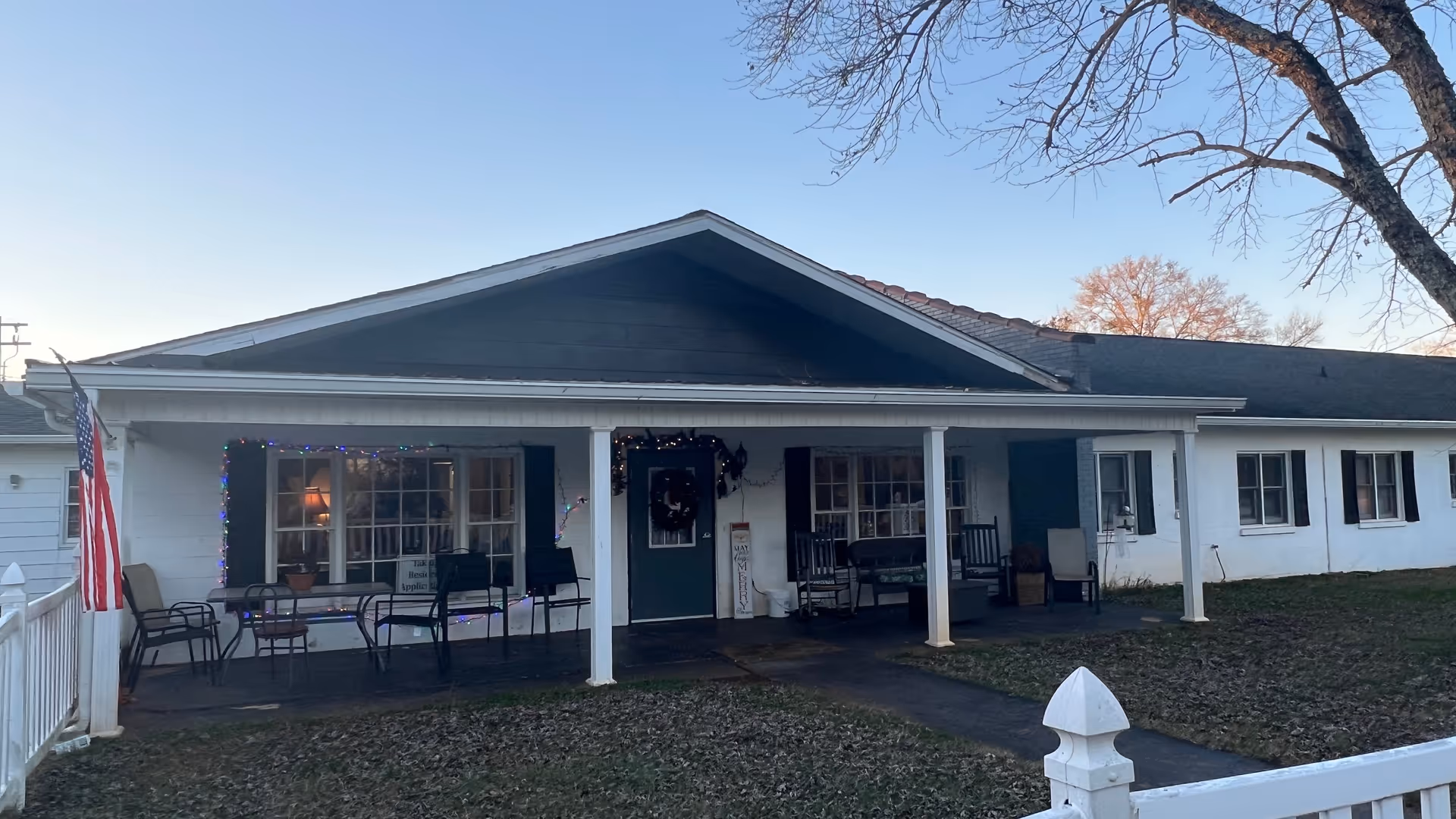 Exterior view of a single-story senior living facility building with a covered porch. The porch has several chairs, a table, and holiday lights around the windows and door. An American flag is displayed on the left side near a white fence. Trees without leaves are visible in the background under a clear sky.
