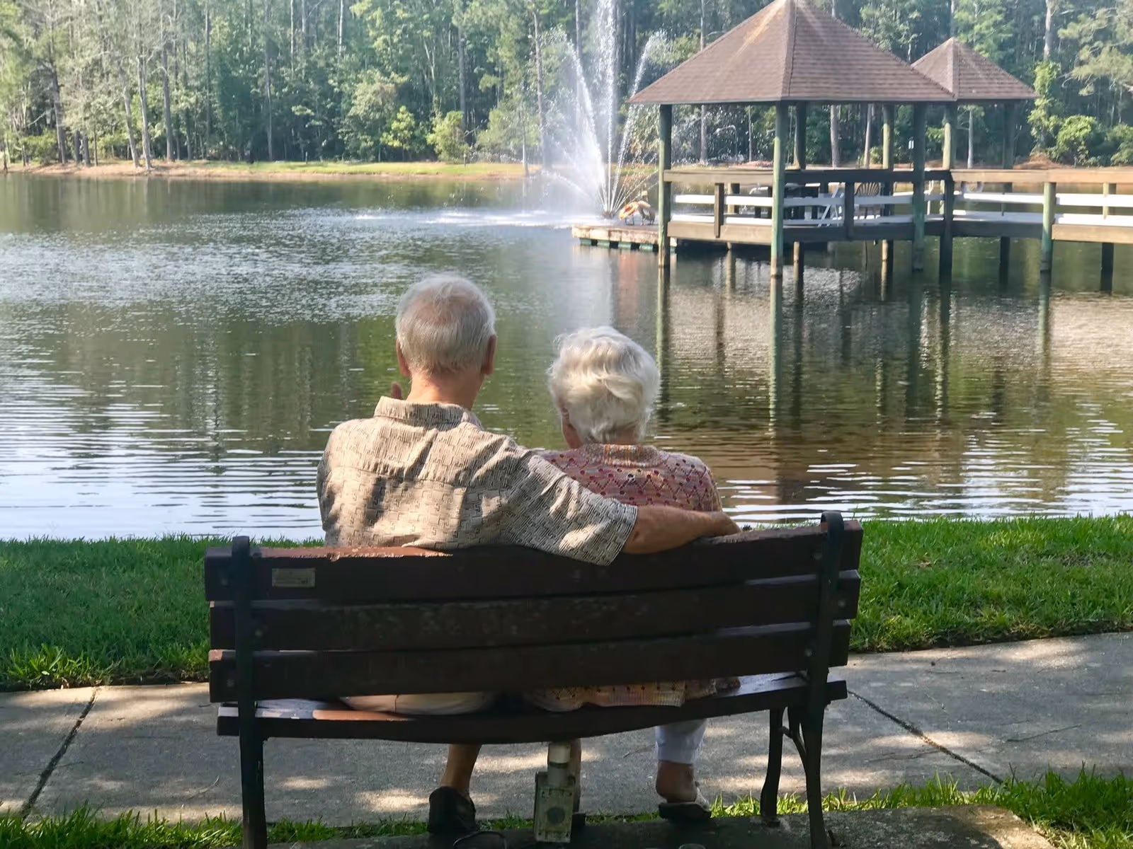 An elderly couple sitting closely on a wooden bench by a lakeside, with the man’s arm around the woman. In the background, there is a gazebo on stilts over the water and a fountain spraying water into the lake, surrounded by trees.