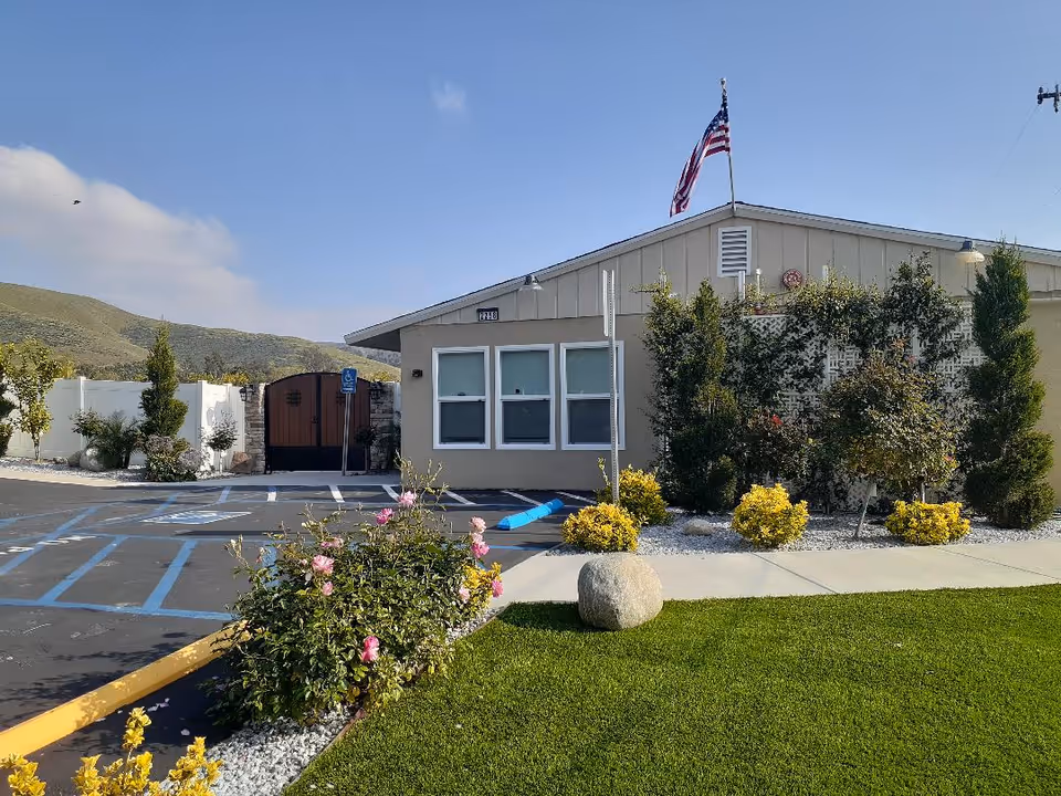 Exterior view of a single-story building with beige walls and three windows. An American flag is flying on a pole on the roof. The building is surrounded by a well-maintained garden with green grass, bushes, and flowers. There is a parking lot with marked handicapped parking spaces in front of the building, and a wooden gate is visible to the left. Hills and a partly cloudy sky are in the background.