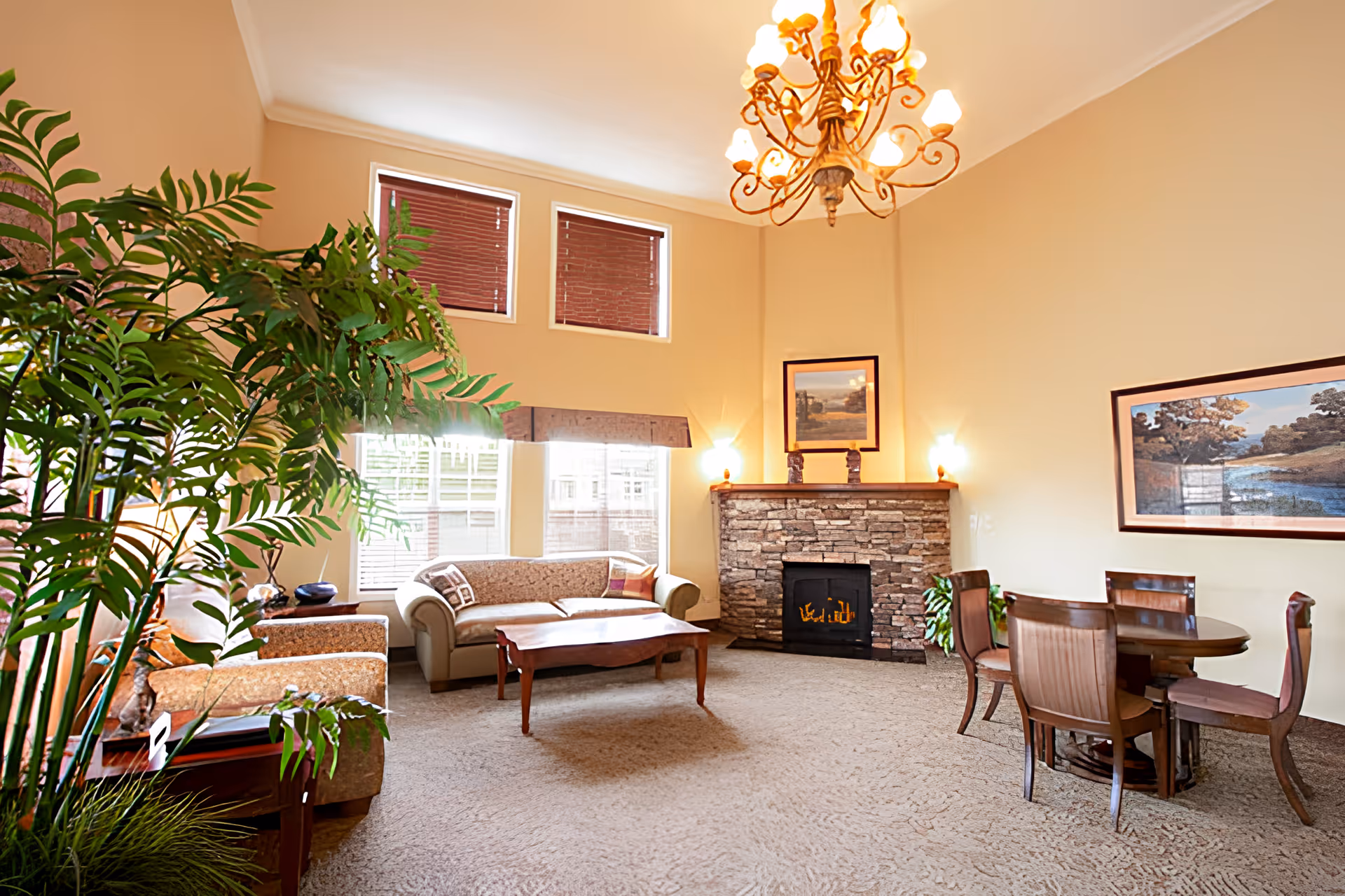 A cozy living room with beige walls and carpet, featuring a stone fireplace with a lit fire, a beige sofa, two armchairs, a wooden coffee table, a round wooden dining table with four chairs, a large green plant, and framed landscape paintings on the walls. The room is illuminated by a chandelier and wall sconces, with natural light coming through three windows with blinds.