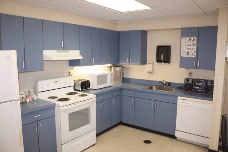 A communal kitchen with blue cabinets and countertops, white appliances including a stove, refrigerator, microwave, and dishwasher.