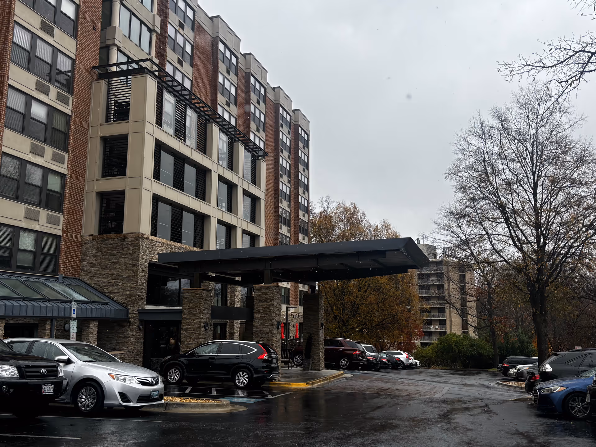 Exterior view of a multi-story senior living facility building with a covered entrance and several parked cars in front on a rainy day. Trees with autumn leaves are visible around the parking area.
