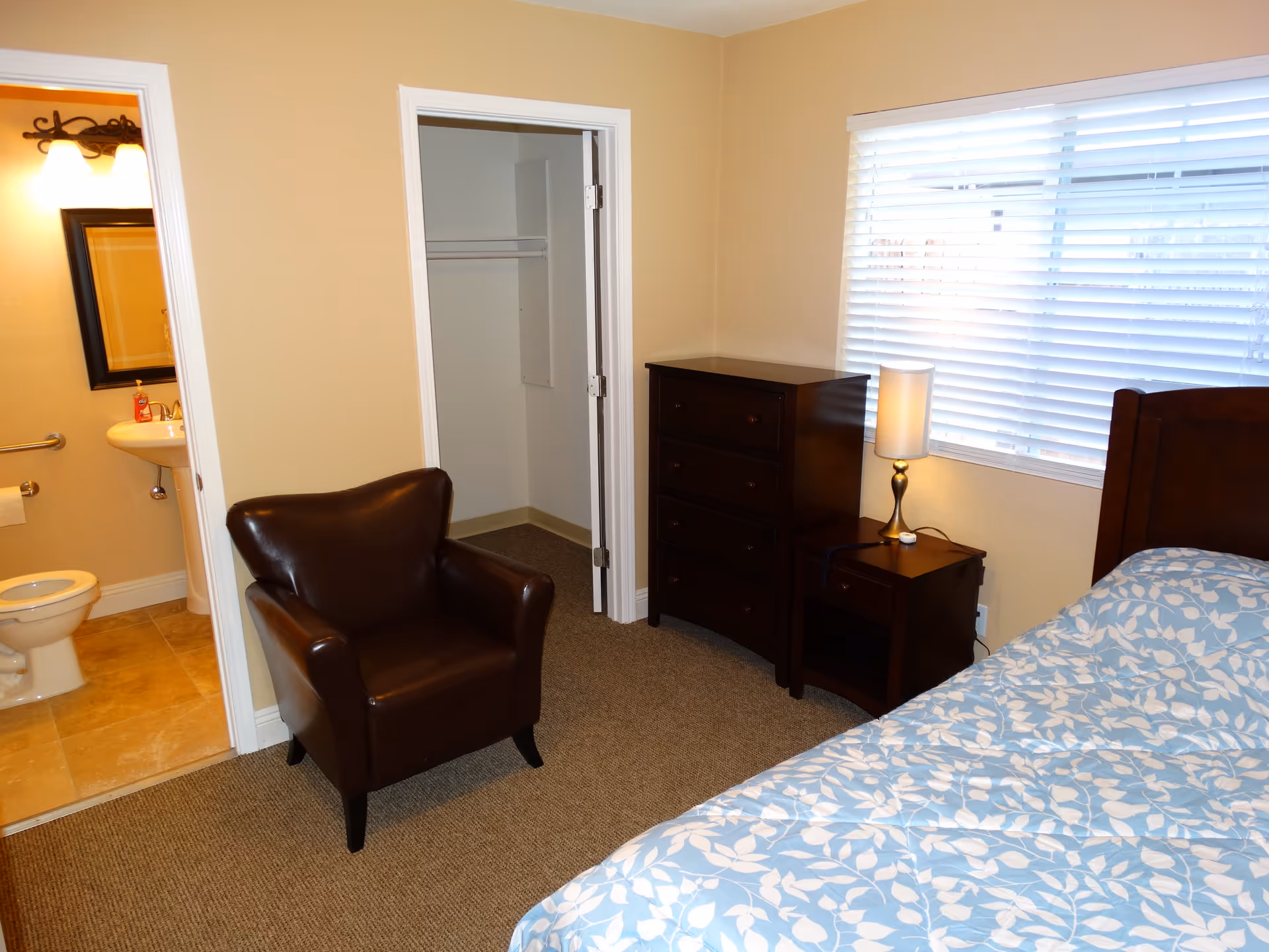 A senior care facility bedroom with a bed covered in a blue and white leaf-patterned comforter, a dark wooden nightstand with a lamp, a matching chest of drawers, a brown leather armchair, a closet with an open door, and an adjacent bathroom with a toilet, sink, and mirror visible.