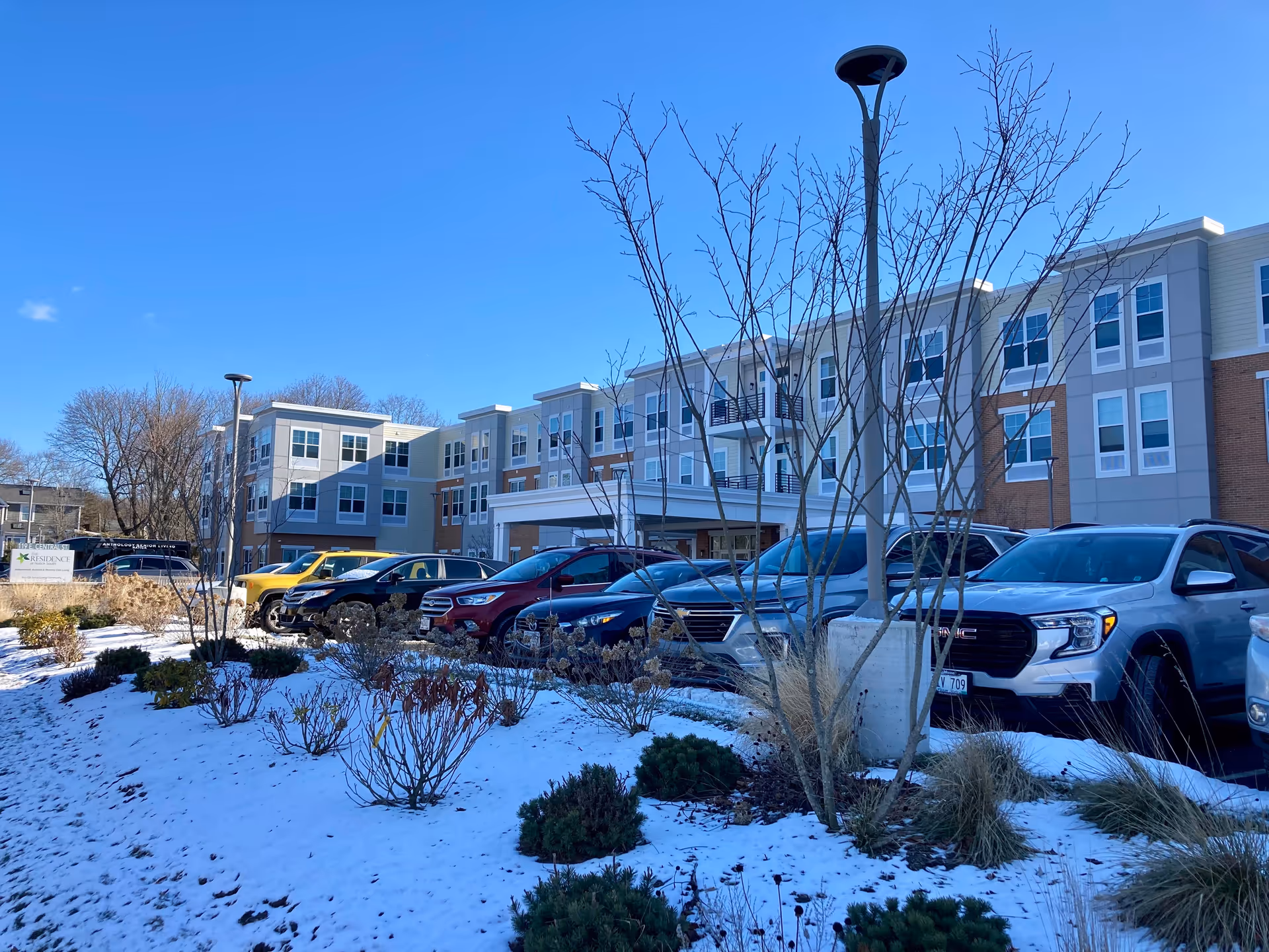 Exterior view of The Residence at Natick South building on a clear winter day with snow on the ground, several parked cars in front, leafless trees, and a blue sky.