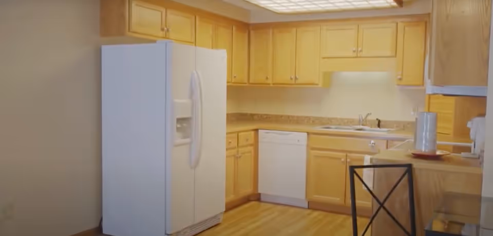 A kitchen with wooden cabinets, a white refrigerator with a water dispenser, a dishwasher, a sink, and a countertop with a pitcher and a plate. There is also a chair with a black metal frame and a wooden floor.