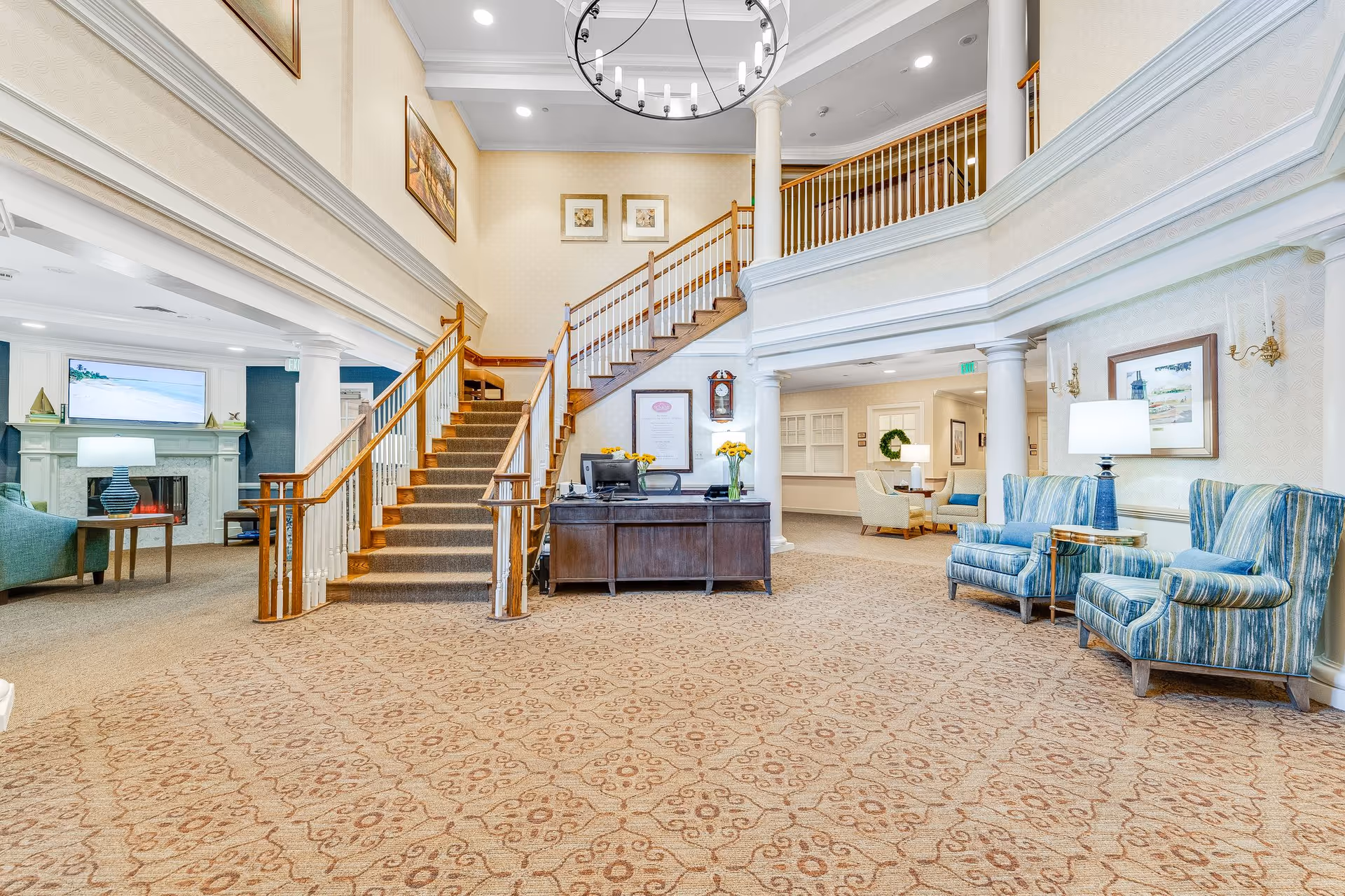Bright senior living facility lobby with a central staircase, reception desk, and seating areas.