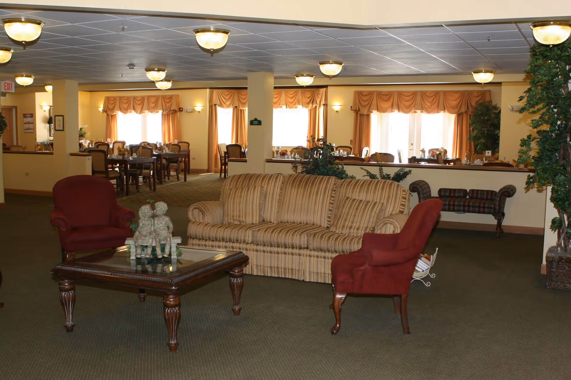A cozy common area in a senior living facility featuring a striped sofa and two red armchairs around a wooden coffee table with a decorative statue. In the background, there is a dining area with multiple tables and chairs, large windows with orange curtains, and ceiling lights illuminating the space.
