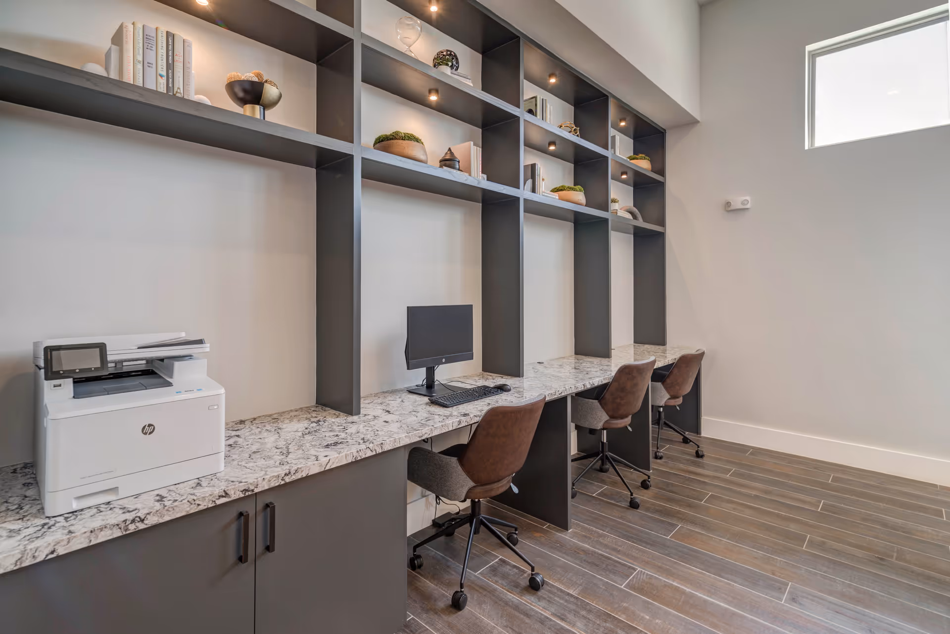 A modern office or workspace area with a long marble countertop, three brown swivel chairs, a computer monitor with keyboard and mouse, a white HP printer, and black shelving units above holding books and decorative items. The room has light-colored walls and a window letting in natural light.