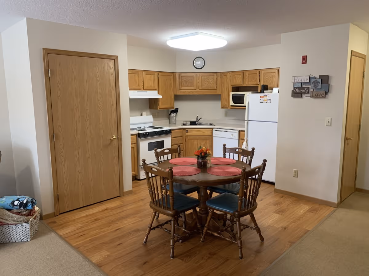 A small kitchen area with wooden cabinets, a white stove, dishwasher, microwave, and refrigerator. In front of the kitchen is a round wooden dining table with four chairs, each with a blue cushion. The table has four red placemats and a small flower arrangement in the center. The floor is a combination of wood and carpet, and there are two wooden doors visible on either side of the kitchen. A wall decoration with the words 'Home', 'Family', and 'Love' is hung on the right wall.