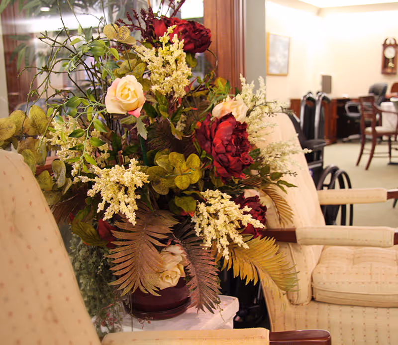 A floral arrangement with red and cream roses, white flowers, and green leaves placed between two beige upholstered armchairs in a room with wooden furniture and a wall clock in the background.