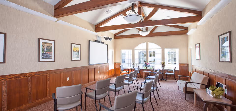 Bright communal meeting room with rows of chairs facing a projection screen, exposed wooden ceiling beams and large arched windows.