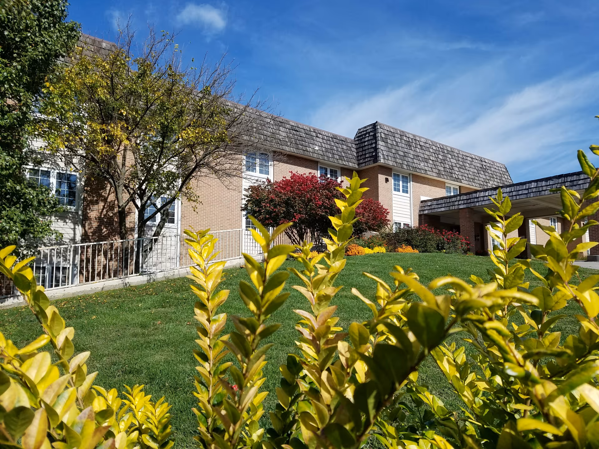 Exterior view of Arbor Court Retirement Community at Alvamar showing a two-story brick building with a shingled roof, surrounded by green grass, yellow leafy bushes in the foreground, and trees with some autumn foliage under a blue sky with light clouds.