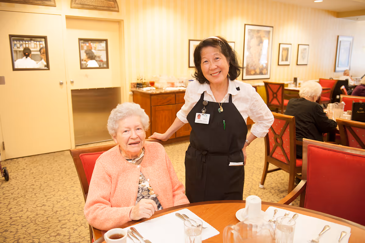 An elderly woman sitting at a dining table with a cup of coffee and utensils, smiling at the camera. A staff member wearing a black apron and name badge stands beside her, also smiling. The background shows other elderly people seated at tables in a warmly lit dining room with framed artwork on the walls.