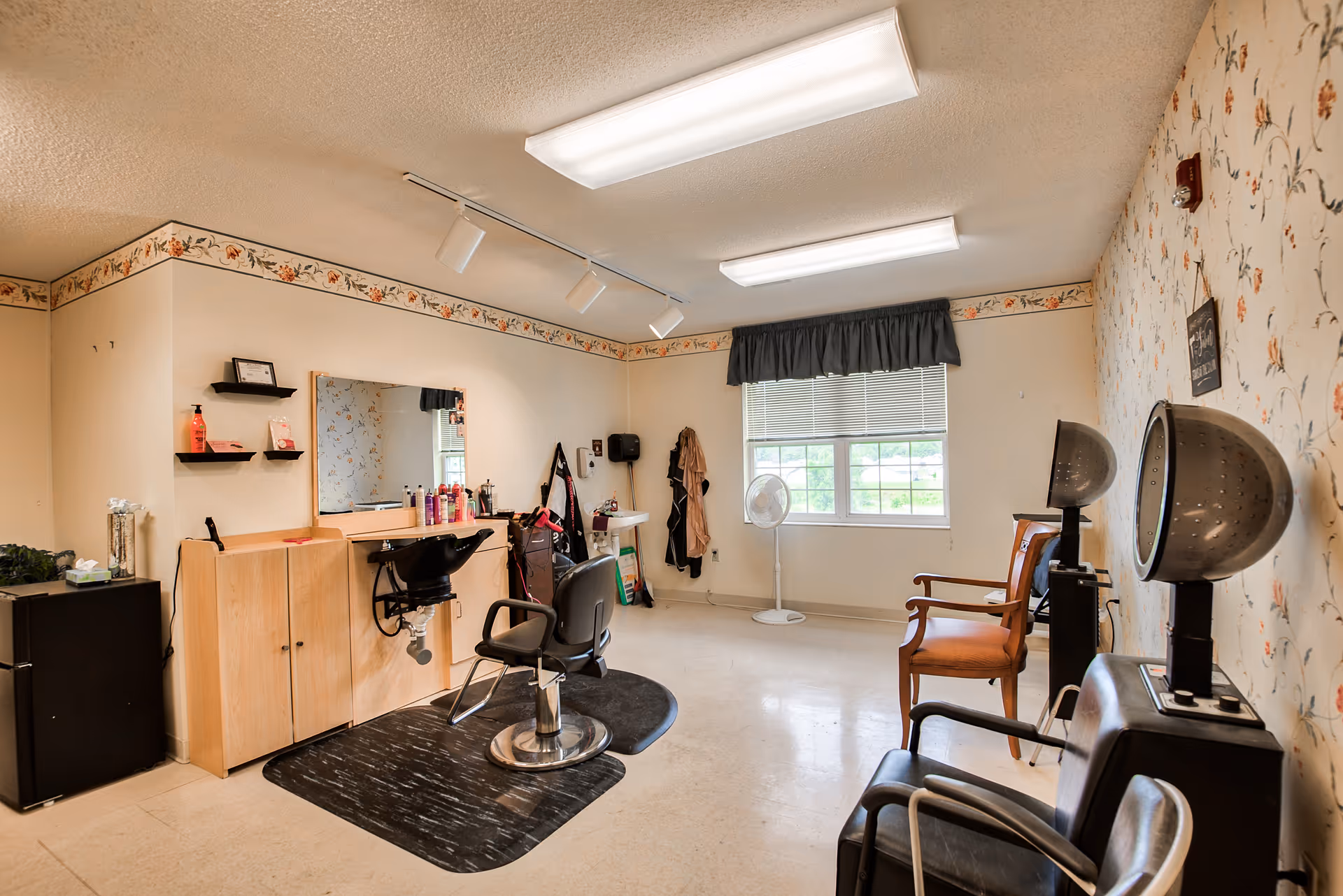 Interior of a small salon room with a hair styling chair in front of a mirror and counter with hair products. There are two hair drying stations with chairs along the right wall, a window with blinds and a fan, and a couple of wooden chairs. The walls have floral wallpaper and a floral border near the ceiling.