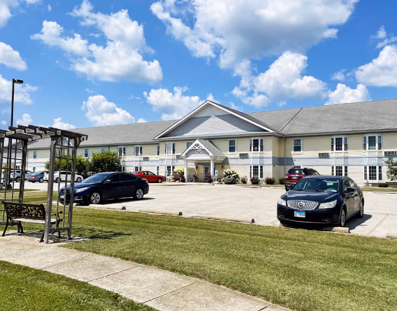 Front exterior of a two-story senior living facility with a parking lot, several cars, a pergola and bench on the lawn under a blue sky.